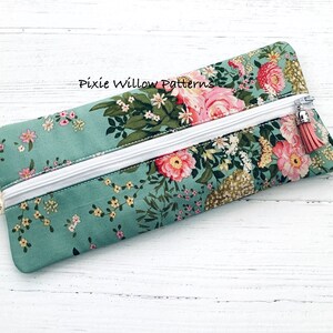 May include: A teal zippered pouch with a floral print featuring pink and white flowers. The pouch has a white zipper and a pink tassel zipper pull. The text "Pixie Willow Patterns" is visible on the pouch.