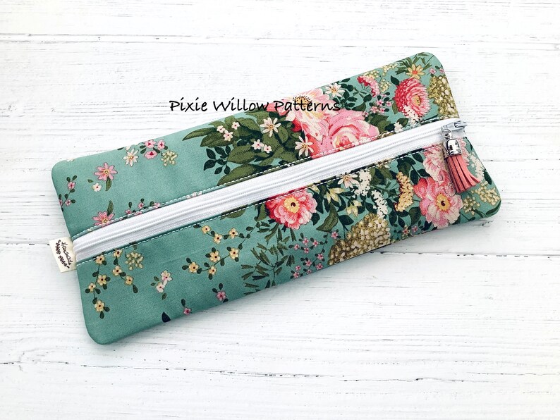 May include: A teal zippered pouch with a floral print featuring pink and white flowers. The pouch has a white zipper and a pink tassel zipper pull. The text "Pixie Willow Patterns" is visible on the pouch.