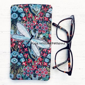 Puede incluir: Un estuche para gafas de tela floral con una libélula azul bordada. El estuche es negro con flores rojas y azules. El estuche está al lado de unas gafas de carey.