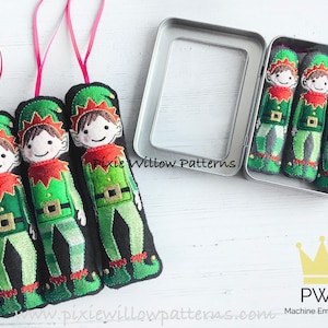May include: Three felt Christmas ornaments shaped like elves, each with a green hat, red collar, and green pants. The elves are in a silver tin with a clear lid. The tin has the text "Pixie Willow Patterns" on it. The ornaments are handmade and perfect for adding a festive touch to your holiday decor.