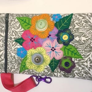 Puede incluir: Una bolsa de tela gris con cremallera y diseño floral. La bolsa tiene un diseño de flores rosa, verde, azul, amarillo y morado con botones en el centro de cada flor. La bolsa tiene una correa rosa con un cierre plateado.