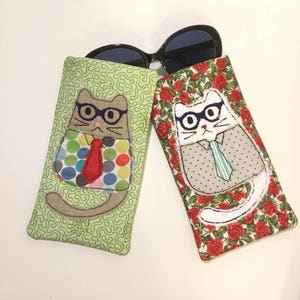 May include: Two cat-themed eyeglass cases. One case is green with a brown cat wearing a red tie and polka dot shirt. The other case is red with white roses and a white cat wearing a blue tie and a gray shirt.