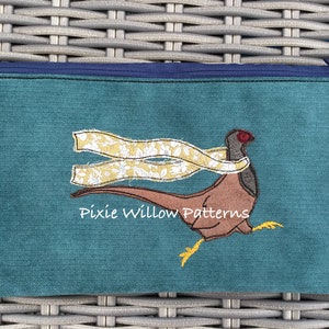 May include: Teal zippered pouch with an embroidered design of a pheasant wearing a yellow and white scarf. The text "Pixie Willow Patterns" is embroidered below the pheasant.