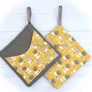 May include: Two yellow and gray quilted pot holders with a floral pattern and a gray loop handle. The pot holders are on a white wooden surface.