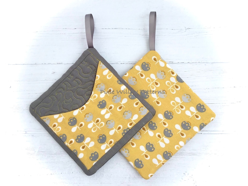 May include: Two yellow and gray quilted pot holders with a floral pattern and a gray loop handle. The pot holders are on a white wooden surface.