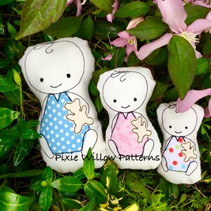 May include: Three handmade plush toys in the shape of babies, each holding a small plush teddy bear. The baby on the left is wearing a blue and white polka dot outfit, the middle baby is wearing a pink and white polka dot outfit, and the baby on the right is wearing a red and white polka dot outfit. The toys are sitting on a bed of green leaves.