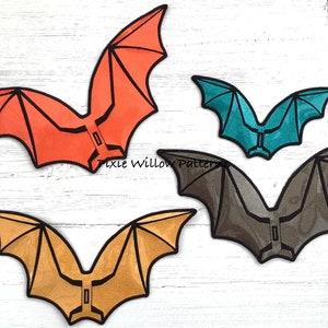 May include: Four colorful bat-shaped decorations in orange, teal, brown, and gold, each with black outlines. The bats have a geometric design and are arranged on a white wooden surface. The text "Pixie Willow Patterns" is visible.