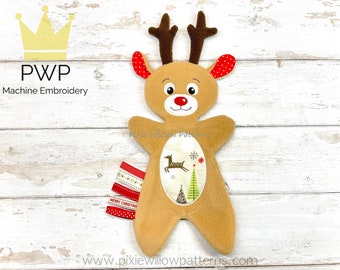 May include: A brown plush reindeer with red ears and antlers. The reindeer has a white embroidered face with a red nose and a white embroidered design on its chest. The design includes a small deer, a snowflake, and two Christmas trees. The reindeer is lying on a white wooden surface.