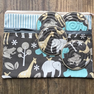 May include: A gray zippered pouch with a blue and white striped lining. The pouch is decorated with a repeating pattern of animals, including giraffes, elephants, frogs, and birds. The pouch contains five round, quilted fabric coasters with animal designs in shades of blue, yellow, and brown.