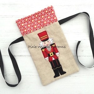 Puede incluir: Una bolsa de tela beige con un lazo de cinta negro. La bolsa presenta una imagen bordada de un soldado de juguete rojo y dorado con una cara blanca y botas negras. El texto "Pixie Willow Patterns" está bordado en la bolsa.