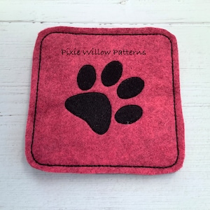 Puede incluir: Un posavasos de fieltro rojo con un dise&ntilde;o de huella de pata negra. El posavasos tiene un borde cosido negro y el texto "Pixie Willow Patterns" est&aacute; impreso en negro.