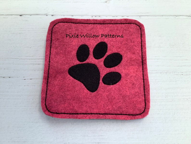 Puede incluir: Un posavasos de fieltro rojo con un dise&ntilde;o de huella de pata negra. El posavasos tiene un borde cosido negro y el texto "Pixie Willow Patterns" est&aacute; impreso en negro.