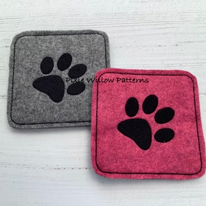 Puede incluir: Dos posavasos de fieltro con huellas de patas negras. El posavasos de la izquierda es gris y el de la derecha es rosa. El texto "Pixie Willow Patterns" es visible en el posavasos rosa.