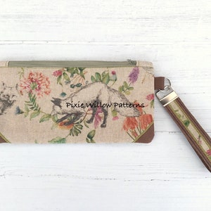 May include: A beige zippered pouch with a floral print featuring a fox, birds, and flowers. The pouch has a brown leather trim and a matching brown strap with a silver clasp.