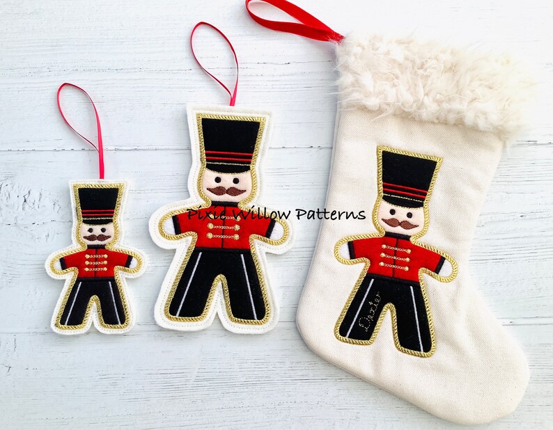 May include: Three felt Christmas ornaments featuring a gingerbread man dressed as a toy soldier. The ornaments are in different sizes. The largest ornament is on a white stocking with a faux fur cuff. The stocking is personalized with the name "Dexter".