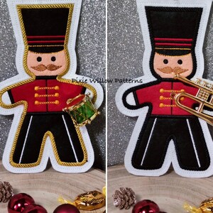 May include: Two felt Christmas ornaments depicting a gingerbread man dressed as a drummer and a trumpeter. The drummer is holding a green and gold drum. The trumpeter is holding a gold trumpet. Both ornaments have black pants, red jackets with gold trim, and black hats with gold trim. The text "Pixie Willow Patterns" is visible in the background.