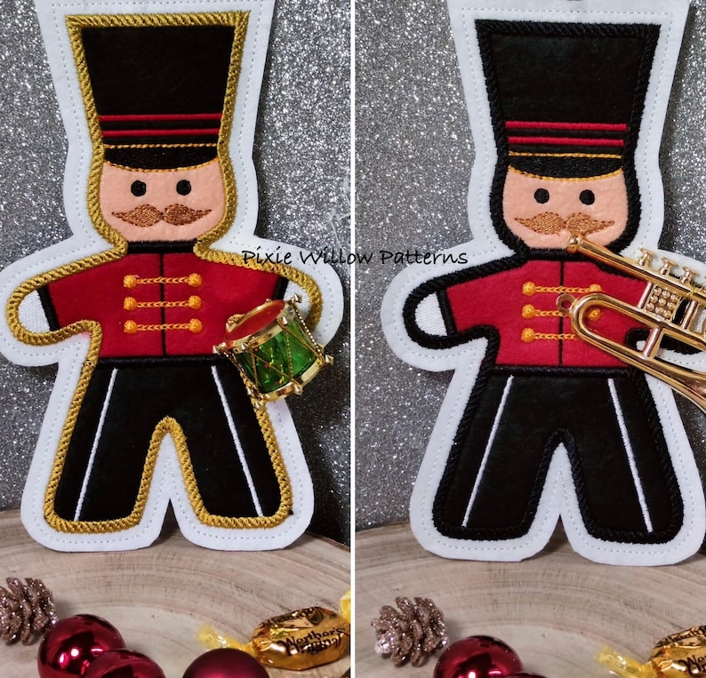 May include: Two felt Christmas ornaments depicting a gingerbread man dressed as a drummer and a trumpeter. The drummer is holding a green and gold drum. The trumpeter is holding a gold trumpet. Both ornaments have black pants, red jackets with gold trim, and black hats with gold trim. The text "Pixie Willow Patterns" is visible in the background.