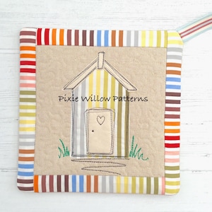 May include: Square, beige fabric pot holder with a striped border in various colors. The center features an embroidered beach hut design with the text "Pixie Willow Patterns". A striped ribbon loop is attached for hanging.
