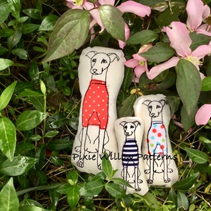 May include: Three handmade plush dog toys in the shape of greyhounds. The largest dog is wearing a red and white polka dot shirt. The middle dog is wearing a blue and white striped shirt. The smallest dog is wearing a green, blue, and white polka dot shirt. The dogs are sitting on a bed of green leaves and pink flowers.