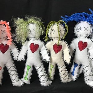 May include: Five handmade voodoo dolls with different coloured yarn hair. Each doll has a red heart, black stitched eyes, and a stitched X for a mouth. The dolls are standing on a black surface.