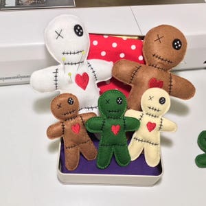 May include: Five felt voodoo dolls in a tin box. The dolls are white, brown, green, and beige. Each doll has a red heart, black eyes, and an embroidered X on its forehead.