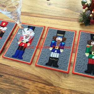 May include: Four quilted coasters featuring different nutcracker designs. Each coaster is rectangular and has a gray background with a red border. The nutcrackers are embroidered in various colors, including red, blue, green, and gold.