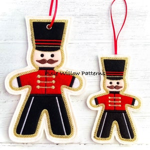 May include: Two felt ornaments shaped like gingerbread men, one larger than the other. Both are dressed as toy soldiers with red jackets, black pants, and gold trim. The larger ornament has a black hat with red stripes and a gold trim. The smaller ornament has a black hat with a gold trim. Both ornaments have a white face with black eyes, a brown mustache, and a red mouth.