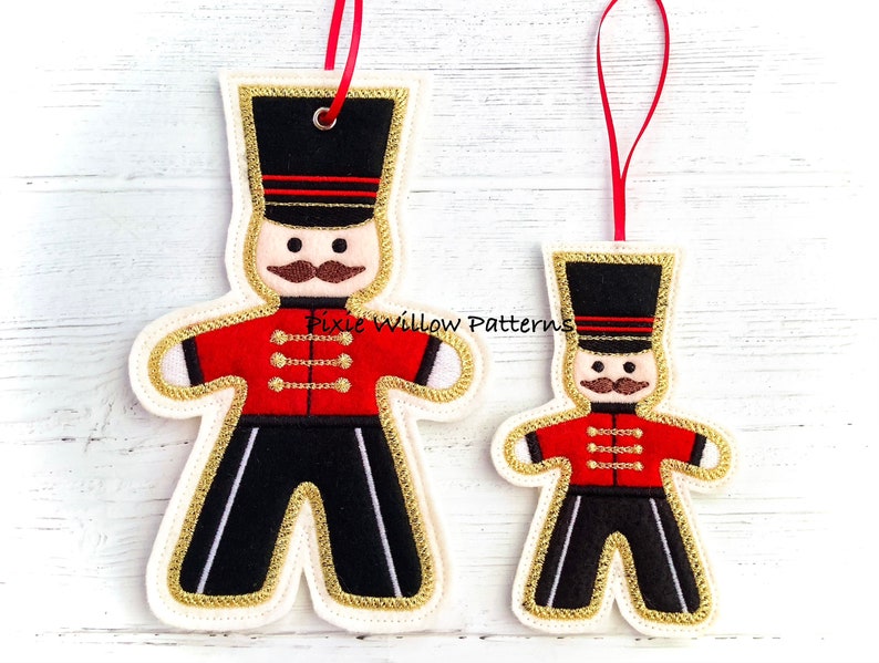 May include: Two felt ornaments shaped like gingerbread men, one larger than the other. Both are dressed as toy soldiers with red jackets, black pants, and gold trim. The larger ornament has a black hat with red stripes and a gold trim. The smaller ornament has a black hat with a gold trim. Both ornaments have a white face with black eyes, a brown mustache, and a red mouth.