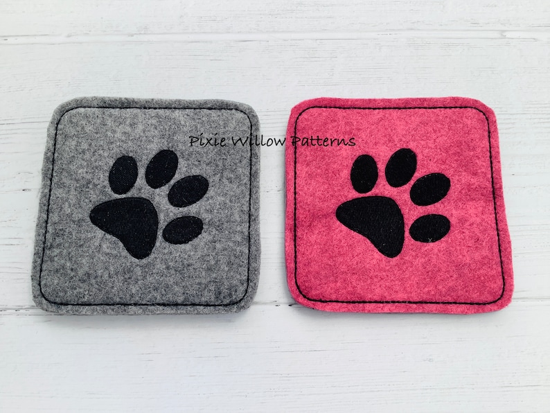 Puede incluir: Dos posavasos de fieltro con huellas de patas negras. El posavasos de la izquierda es gris y el de la derecha es rosa. El texto "Pixie Willow Patterns" est&aacute; impreso en los posavasos.