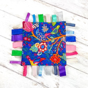 May include: A blue fabric baby sensory toy with colorful ribbons and a floral pattern. The fabric features two birds and the text "Pixie Willow Patterns".