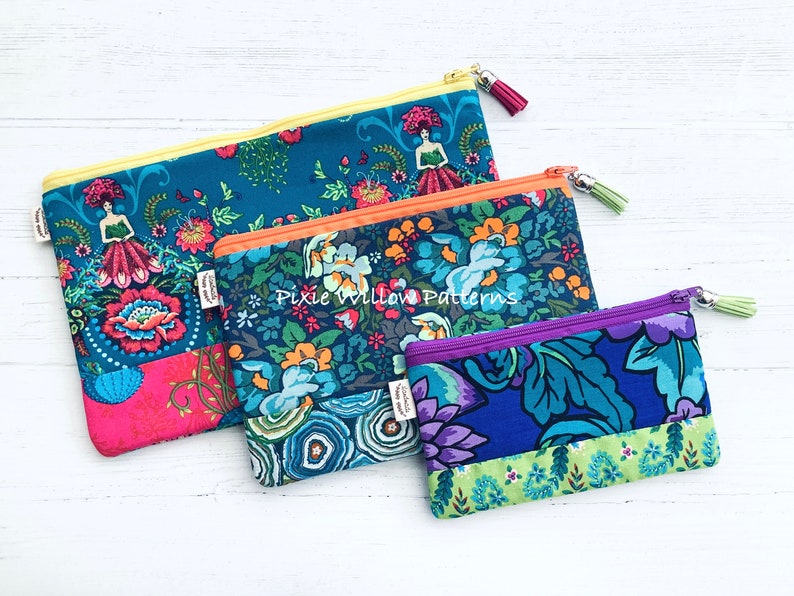 Op de afbeelding: Drie ritszakjes met bloemenpatronen in tinten blauw, groen, paars, oranje en roze. De zakjes zijn gevoerd met een contrasterende stof en hebben een ritssluiting met een kwast. Het grootste zakje heeft een gele rits en de tekst "Pixie Willow Patterns" gedrukt op de voorkant.