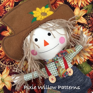 Pode incluir: Uma boneca de espantalho feita à mão com um chapéu de feltro castanho, um girassol amarelo bordado no chapéu, uma camisa xadrez, suspensórios vermelhos e um macacão de ganga azul. A boneca está deitada sobre um fundo de tecido com um padrão de girassol.