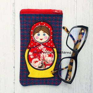 May include: A red, yellow, and white embroidered Russian nesting doll design on a blue and red fabric eyeglass case with a zipper. The case is lying on a white wooden surface next to a pair of tortoiseshell eyeglasses.