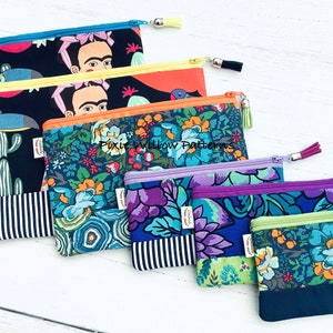 Op de afbeelding: Vijf kleurrijke ritszakjes met verschillende patronen. De zakjes zijn in een rij van groot naar klein gerangschikt. Het grootste zakje heeft een zwarte achtergrond met een kleurrijk beeld van Frida Kahlo en een toekan. De andere zakjes hebben bloemmotieven in tinten blauw, paars en groen. De zakjes hebben een gestreepte stof aan de onderkant en een kwast rits.