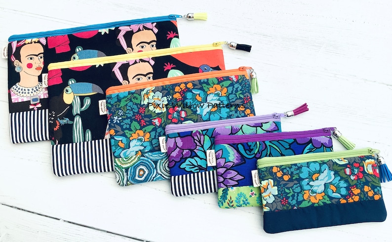 Op de afbeelding: Vijf kleurrijke ritszakjes met verschillende patronen. De zakjes zijn in een rij van groot naar klein gerangschikt. Het grootste zakje heeft een zwarte achtergrond met een kleurrijk beeld van Frida Kahlo en een toekan. De andere zakjes hebben bloemmotieven in tinten blauw, paars en groen. De zakjes hebben een gestreepte stof aan de onderkant en een kwast rits.