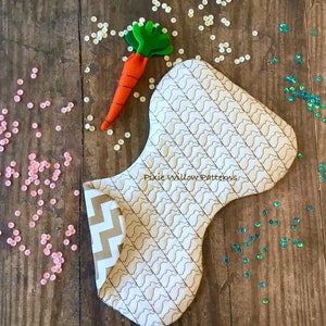 May include: A white fabric bunny-shaped pillow with a brown zig-zag pattern on the side. The pillow is quilted with a brown pattern of bunny silhouettes. The pillow is surrounded by pink and green sequins on a wooden surface. The text "Pixie Willow Patterns" is embroidered on the pillow.