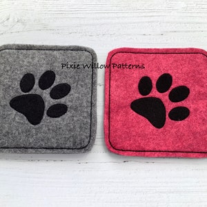 Puede incluir: Dos posavasos cuadrados de fieltro con bordado de huella de pata negra. El posavasos de la izquierda es gris y el de la derecha es rosa. El texto "Pixie Willow Patterns" est&aacute; bordado en los posavasos.
