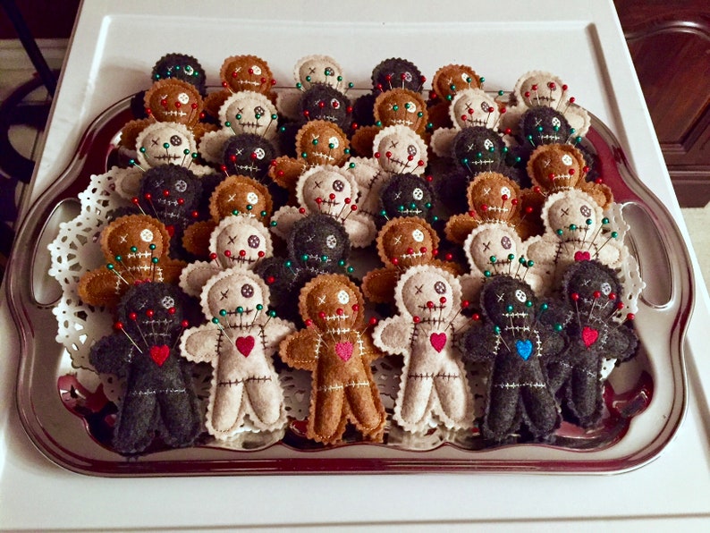 May include: A silver tray filled with felt voodoo dolls. The dolls are in various colors, including black, brown, white, and gray. Each doll has a red, blue, or green heart sewn on the front and is decorated with pins.