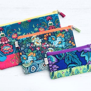 Op de afbeelding: Drie ritszakjes met bloemenpatronen in tinten blauw, groen en paars. De zakjes zijn gevoerd met een contrasterende stof en hebben een ritssluiting met een kwastje. Het grootste zakje heeft een gele rits en een roze en blauwe bloemenprint. Het middelste zakje heeft een oranje rits en een blauwe en groene bloemenprint. Het kleinste zakje heeft een paarse rits en een blauwe en groene bloemenprint. De zakjes zijn gelabeld met "Pixie Willow Patterns".