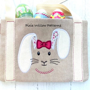 May include: A beige Easter tote bag with a bunny face applique and the text "Pixie Willow Patterns." The bunny has white ears with floral print lining, a red bow, and embroidered details. The bag contains three decorated Easter eggs.