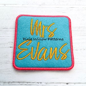 May include: A blue felt square with a pink border, featuring the embroidered text "Mrs. Evans" in gold thread. The text "Pixie Willow Patterns" is also embroidered in gold thread below the name.
