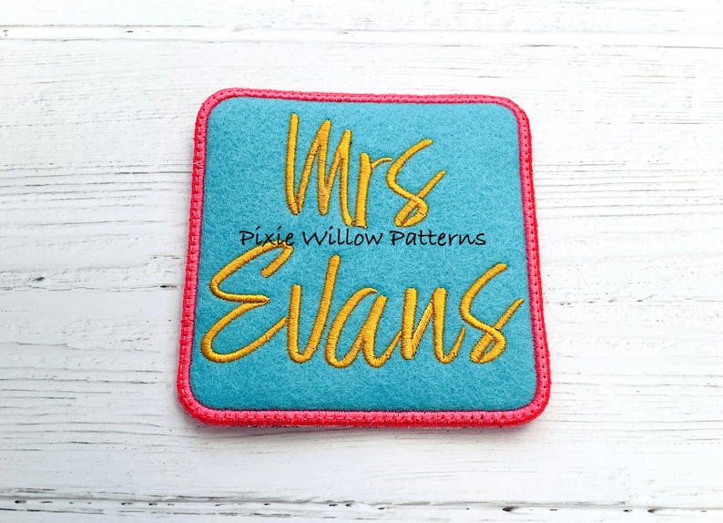 May include: A blue felt square with a pink border, featuring the embroidered text "Mrs. Evans" in gold thread. The text "Pixie Willow Patterns" is also embroidered in gold thread below the name.