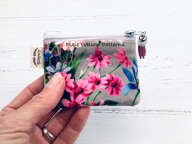 May include: A small floral print zipper pouch with a pink tassel. The pouch has a white zipper and a silver charm. The pouch is handmade and says "Pixie Willow Patterns" on the front.