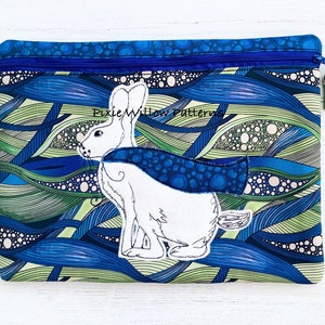 Könnte beinhalten: Eine blaue und grüne Reißverschlusstasche mit einem weißen Kaninchen, das einen blauen Umhang mit weißen Punkten trägt. Die Tasche hat einen blauen Reißverschluss und einen Quasten-Zipper. Der Text "Pixie Willow Patterns" ist auf der Tasche aufgedruckt.