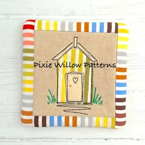 May include: A yellow and green striped beach hut with a red heart on the door, embroidered on a beige fabric square with a colorful striped border. The text "Pixie Willow Patterns" is embroidered on the fabric.