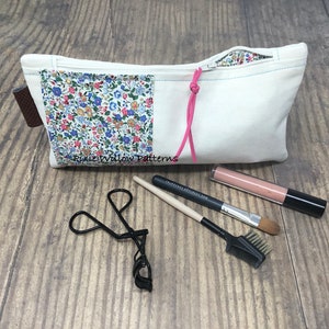 May include: A white zippered pouch with a floral fabric lining and a pink drawstring. The pouch is lying on a wooden surface with a black eyelash curler, a makeup brush, and a tube of lip gloss.