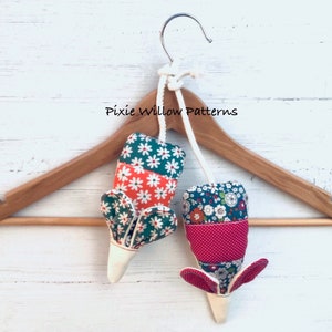 Puede incluir: Dos adornos colgantes de tela con forma de ratón, uno con tela verde, blanca y naranja y el otro con tela roja, blanca y azul. Los adornos cuelgan de una percha de madera con el texto "Pixie Willow Patterns" impreso en ella.