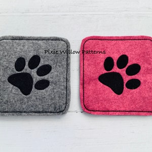 Puede incluir: Dos posavasos de fieltro con huellas de patas negras. El posavasos de la izquierda es gris y el de la derecha es rosa. El texto "Pixie Willow Patterns" est&aacute; impreso en los posavasos.