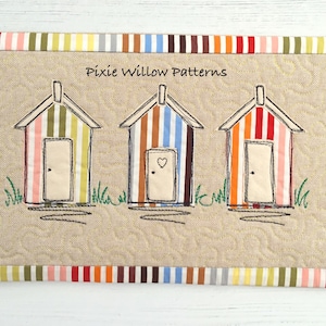 Puede incluir: Una pared de tela beige con un borde de rayas de colores. El centro de la pared presenta tres casas de playa bordadas con puertas y un corazón en una de las puertas. El texto "Pixie Willow Patterns" está bordado sobre las casas.