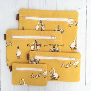 Może przedstawiać: Cztery żółte saszetki z zamkiem błyskawicznym, białym zamkiem i nadrukiem kaczki. Saszetki są ułożone jedna na drugiej, z największą saszetką na dole i najmniejszą na górze. Saszetki są wykonane z miękkiej, wytrzymałej tkaniny i mają zamek błyskawiczny. Saszetki idealnie nadają się do przechowywania drobnych przedmiotów, takich jak kosmetyki, biżuteria lub elektronika.